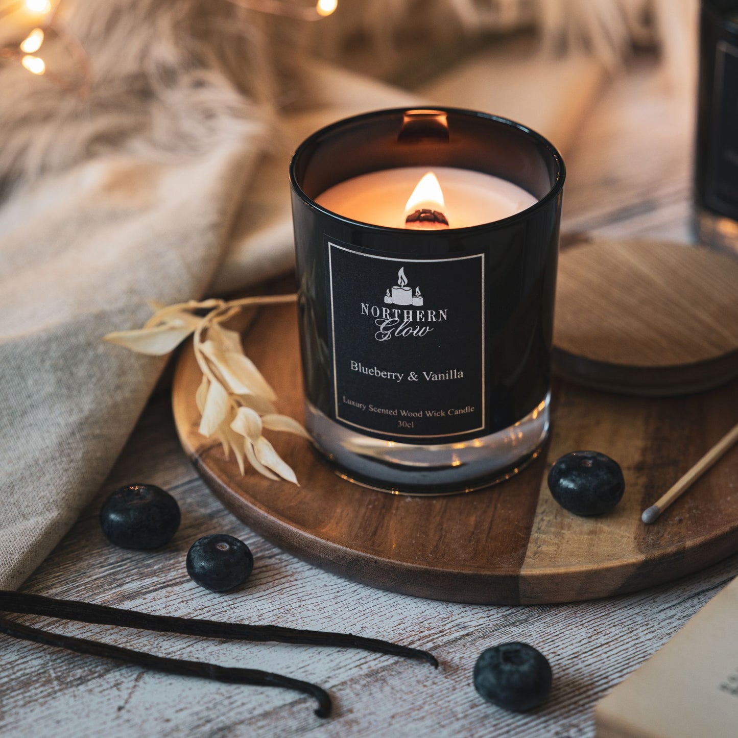 A black jar with the wood wick lit and a nice flame burning. It has blueberries in front of the jar with some vanilla pods.
