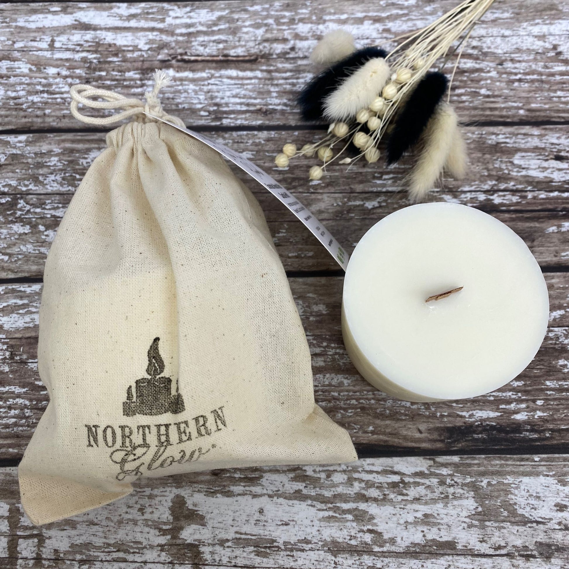 A refillable wax candle ready to put into a jar. It has a wood wick and the refill candle is in a white cotton bag, tied at the top. It has a black stamp with Northern Glow logo, on the cotton bag. It has a white tag tied to the bag with product usage details and ingredients.