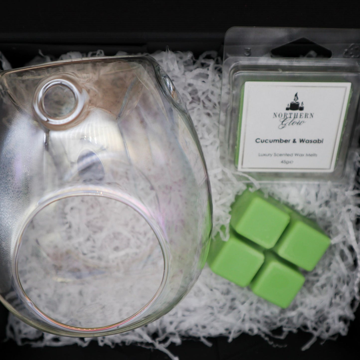 Clear pearl colour glass wax melt burner in a black gift box with a green wax melt in a square shape.