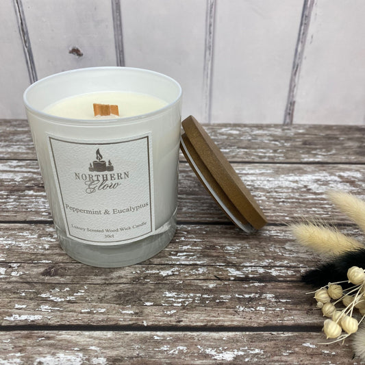 A white glass candle jar with a wooden lid leaning against the jar. A white label with logo and Peppermint and Eucalyptus near the bottom of label. It ahs a luxury feel.