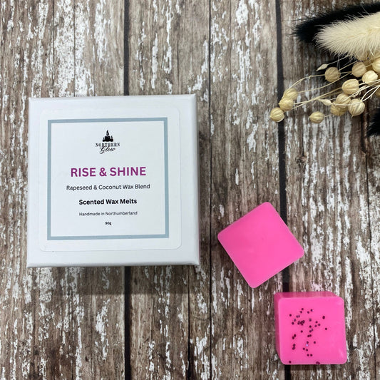 Rise & Shine wax melt is a magenta colour and each melt is a square shape in a white box. There are eight individual wax melts in the white box. The label is white with a silver border. The logo is at the top with 'Rise & Shine' in pink writing in the centre.