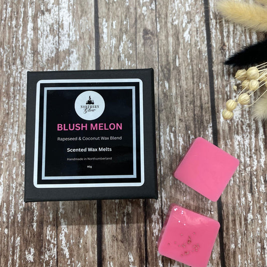 Blush Melon scented wax melts packaging on a wooden surface. Wax Melts are in a small black box with the name Blush Melon in pink writing.