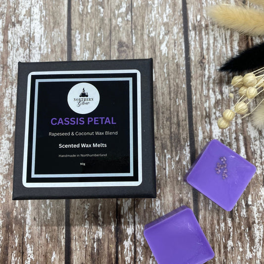 A box of Cassis Petal wax melts alongside two individual purple wax melt cubes, presented on a wooden surface with decorative dry plants in the background.