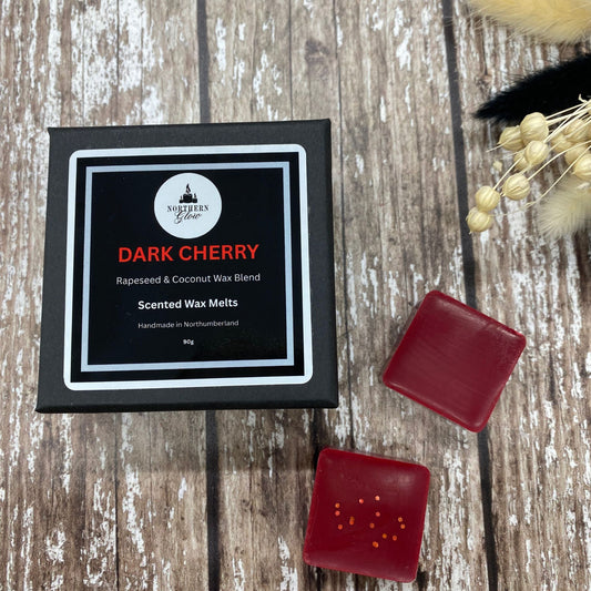 A package of Dark Cherry scented wax melts from Northern Glow, with two red square wax melts placed outside the package on a wooden surface, accompanied by decorative dried flowers.