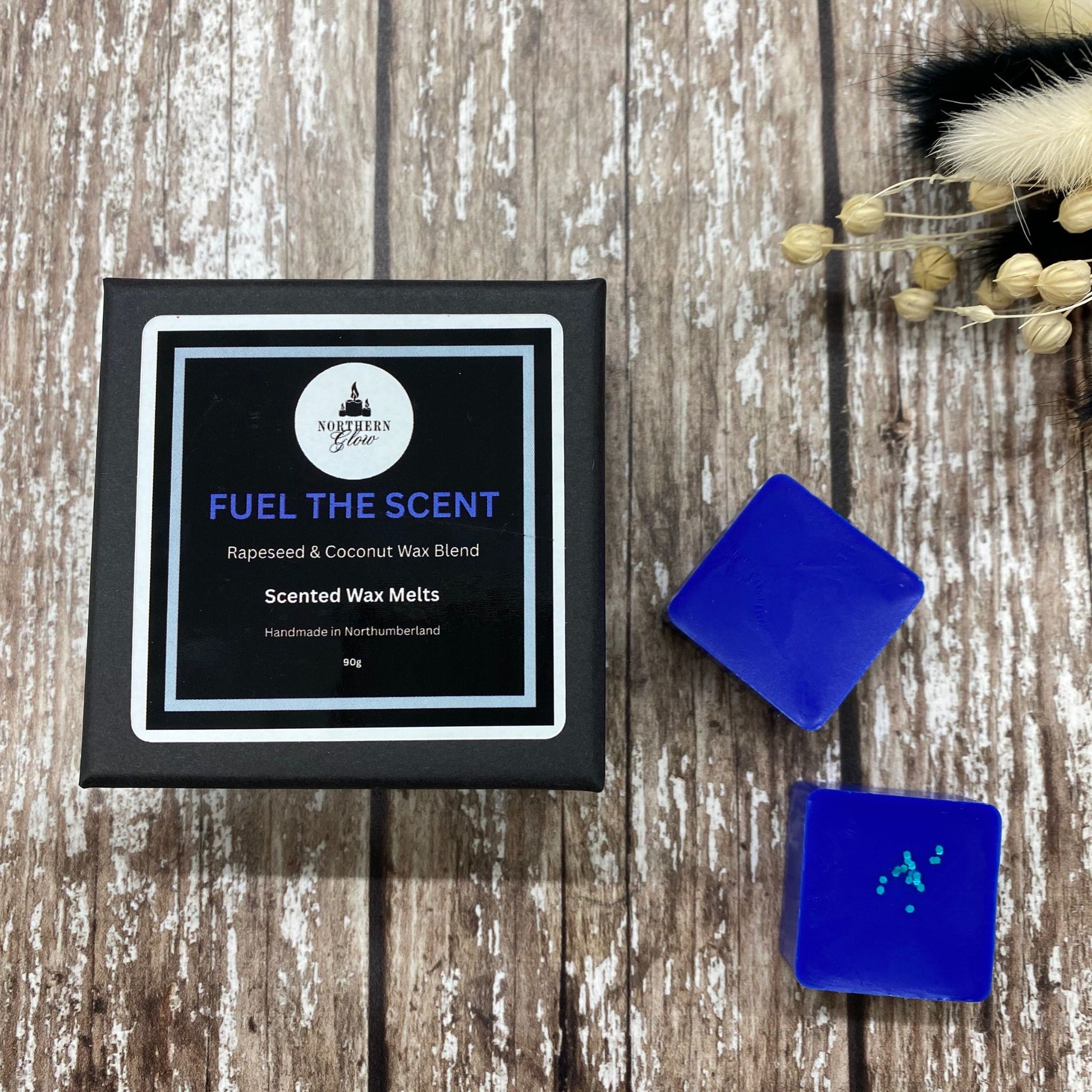 A black square box with two square dark blue wax melts next to the box, on a wood background. The logo is on the label with the fragrance Fuel the Scent in dark blue writing.