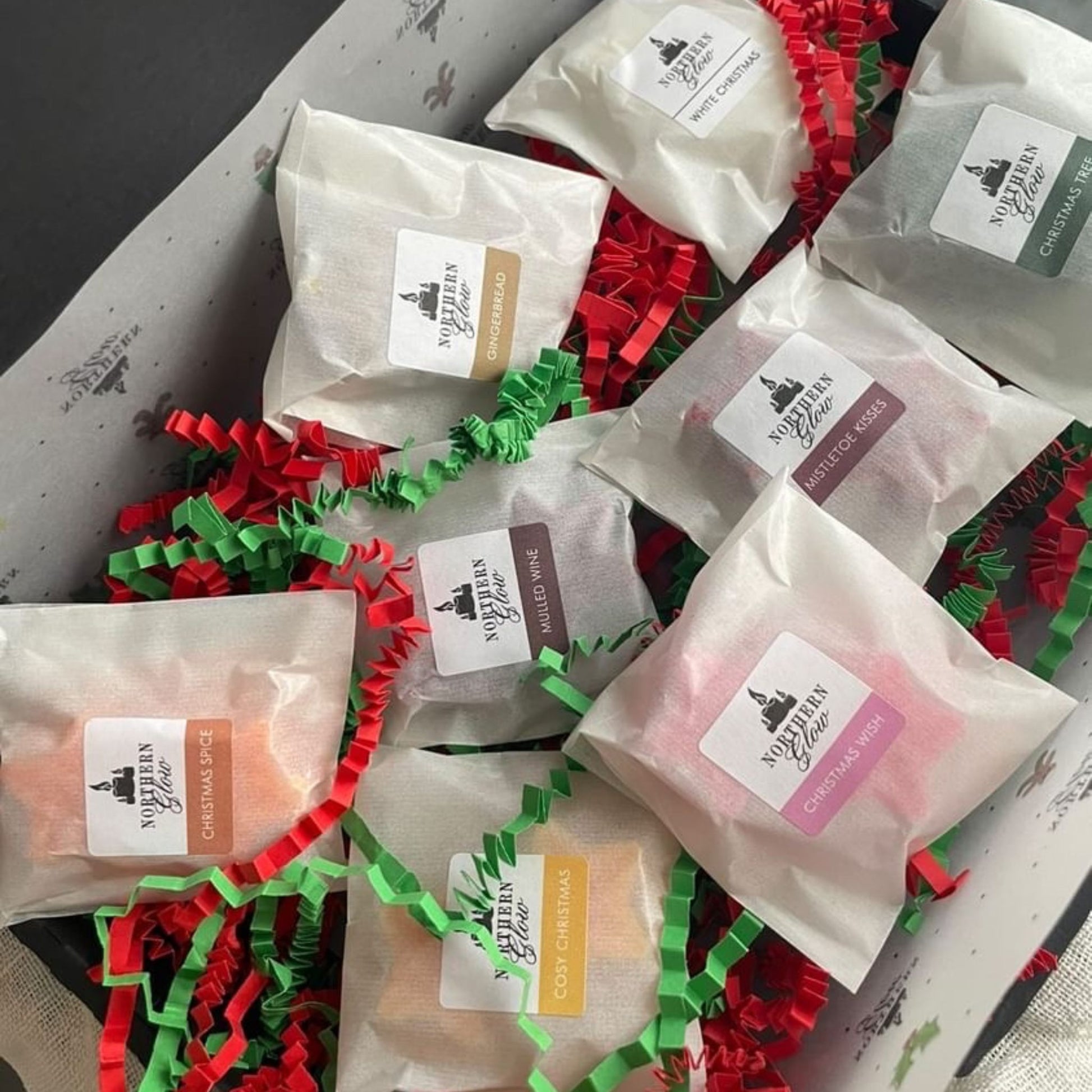 The Christams Wax Melt sample box is in a black box with eight different fragrance. Each fragrance is in a glassine bag with two wax melt stars in each bag. There is green and red shredded paper in the box.