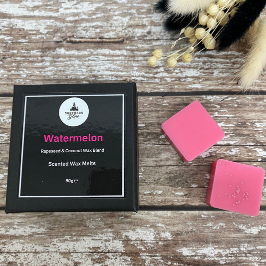A small lack box with a black label with the word Watermelon on it, in pink writing. It has the logo at the top. There are 2 wax melt squares at the side of the box, in pink colour.