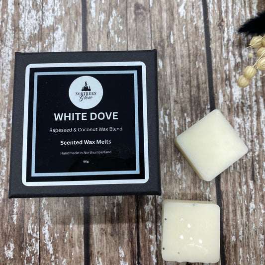 A package labeled 'White Dove' with two square wax melts beside it, placed on a wooden surface with dried plants in the background.
